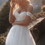Blond woman in wedding gown featuring off the shoulder straps, optional sleeves, intricate lace and bead details and sparkling tulle